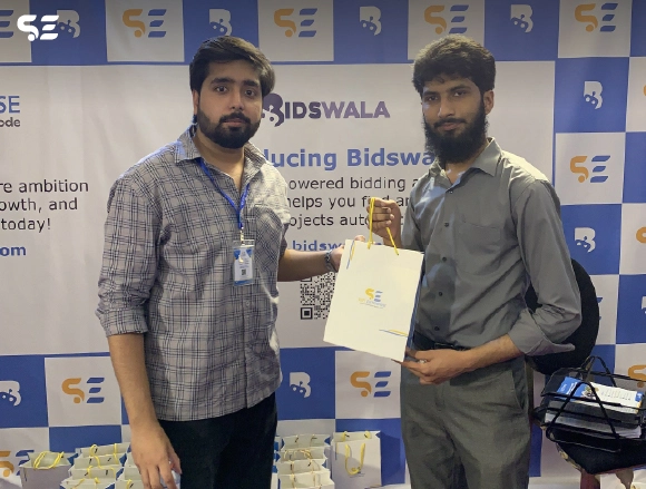 Two male employees posing together in front of Bidswala banner
